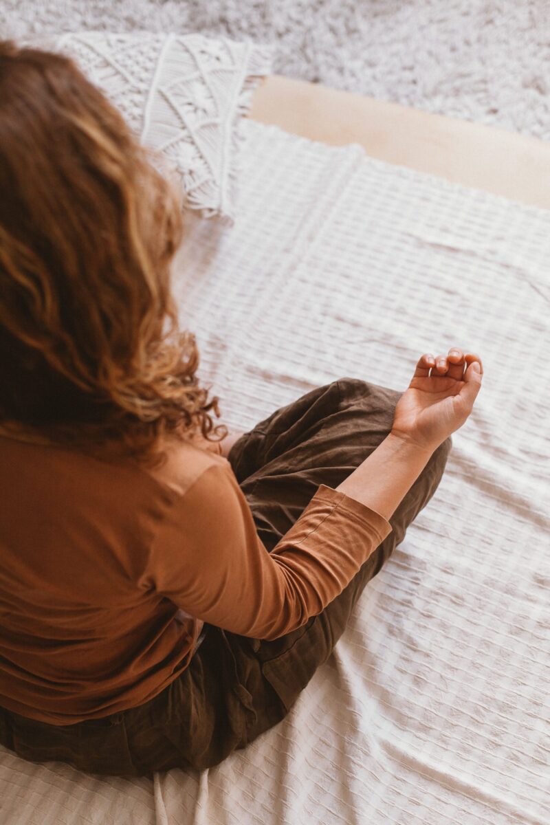 A woman in brown attire meditates on a soft surface indoors, promoting peace and mindfulness.